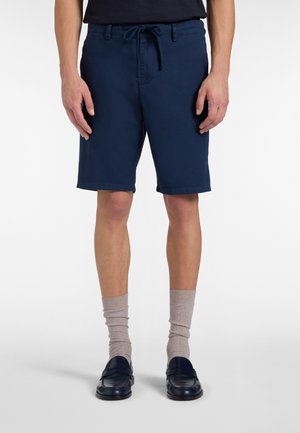 Man wearing navy blue drawstring shorts, beige socks, and black loafers, standing with arms relaxed by sides.