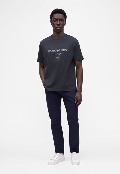Νέος άνδρας που φοράει μαύρο t-shirt Emporio Armani, σκούρο παντελόνι και άσπρα αθλητικά παπούτσια στέκεται μπροστά σε μια απλή λευκή επιφάνεια.
