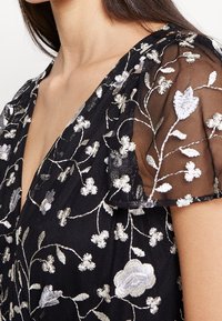 Černé šaty s květinovou výšivkou ve stříbrné a bílé barvě, s jemným vyšívaným pláštěm na rukávech. Měkká textura a výstřih do V.