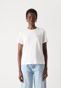 T-shirt bianco in cotone con colletto rotondo e maniche corte, con stampa di testo multicolore ben visibile sul davanti. Abbinato a jeans azzurri chiari.