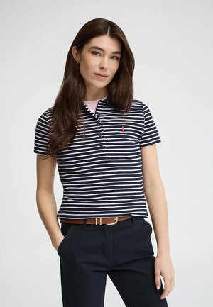 Jeune femme aux longs cheveux bruns portant un polo rayé bleu marine et blanc, un pantalon noir et une ceinture marron, posant avec les mains dans les poches.
