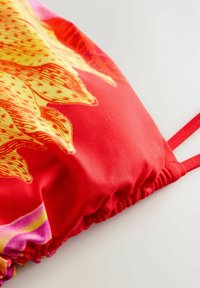 Tissu rouge avec imprimé de fleurs jaunes et oranges, avec des détails froncés et de fines bretelles rouges sur fond blanc.