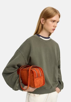 Młoda kobieta w szarej bluzie trzyma małą prostokątną pomarańczową zamszową torebkę z podwójnymi suwakami i paskami na ramię.