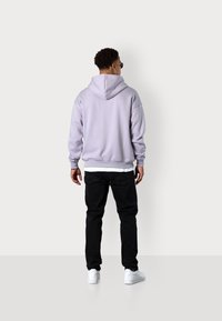 Light lavender hooded sweatshirt with ribbed cuffs and hem, black jeans, and white sneakers, viewed from the back against a neutral background.