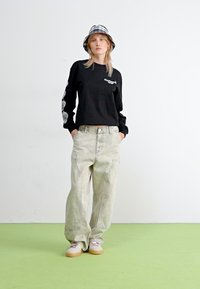 Svart langermet skjorte med hvit grafikk, sammen med lysegrå baggy bukser og beigefargede sneakers på et grønt gulv. Bøttehatt med rutete mønster.