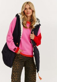 Frau mit blonden Haaren, die einen rosa und roten Pullover, eine schwarze Steppweste mit weißem Fleecefutter und eine Hose mit Leopardenmuster trägt, posiert mit einer Hand in der Tasche.