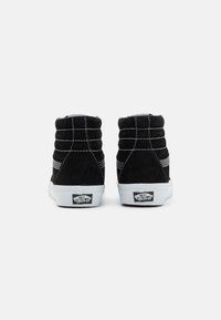 Čierne semišové tenisky s vysokým strihom, biele švy a gumové podrážky. Obsahujú polstrovaný golier a logo Vans na päte.
