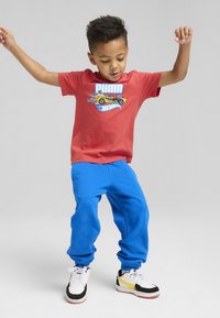 Camiseta de algodón roja con el texto "PUMA" y un gráfico de coche, combinada con pantalones de chándal azules. Incluye zapatillas blancas con acentos negros y detalles amarillos.