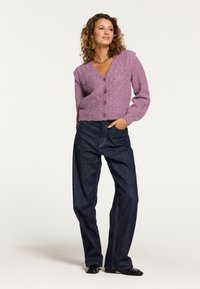 Fialový pletený cardigan s výstřihem do V a třemi knoflíky, zkombinovaný s tmavými džíny s vysokým pasem. Kadeřavé vlasy a minimální doplňky.