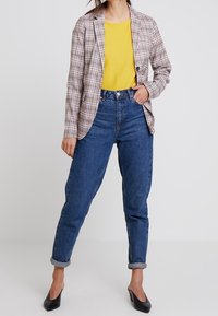 Hnědo-bílý kostkovaný blazer přes žlutý top, doplněný o ohrnuté tmavě modré džíny a černé špičaté podpatky.