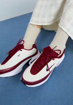 Pieds portant des baskets Nike blanches et bordeaux avec des chaussettes crème côtelées, sur un sol moquetté bleu, avec un pantalon blanc texturé visible.