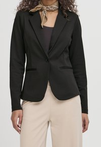 Blazer noir en tissu lisse avec une fermeture à un seul bouton, des revers structurés et des poches avant. Associé à un pantalon beige.