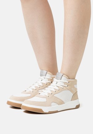 Jambes portant des baskets montantes beige et blanches avec des lacets blancs, logo "BOSS" sur la languette et le côté, debout sur un fond clair.