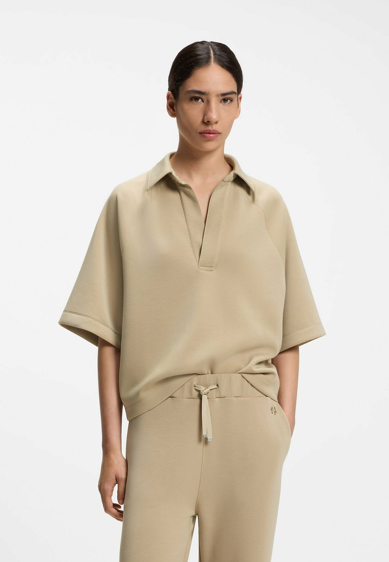 Femme aux cheveux foncés portant un haut beige à manches courtes col polo et un pantalon assorti à cordon, debout devant un fond blanc uni.