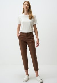 T-shirt bianca a maniche corte abbinata a pantaloni marroni a gamba affusolata con tasche laterali e una trama liscia, completata da sneakers bianche.