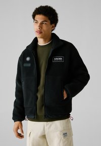 Sort fleecejakke med høj krave, to frontlommer og broderede logoer. Båret over en olivengrøn sweatshirt og lysebeige bukser.