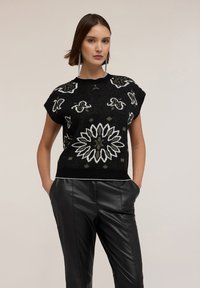 Suéter negro de punto con mangas cortas, que presenta intrincados patrones florales en blanco y gris, acompañado de unos elegantes pantalones de cuero negros.