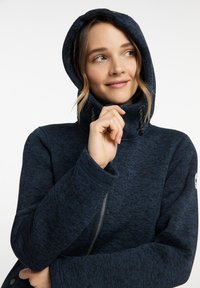 Jūras sarkanā kapuce izgatavota no tekstūrēta auduma, ar rāvējslēdzēju, regulējamu kapuci ar auklām un logotipu uz piedurknes.