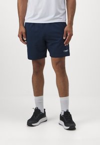 Mand iført marineblå træningsshorts, hvide sokker, sorte sneakers og en hvid skjorte, der står mod en ensfarvet baggrund.