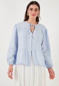 Blusa azzurro chiaro con abbottonatura frontale, collo a fiocco e maniche a palloncino. Realizzata in un tessuto leggero e stropicciato con orlo peplum.
