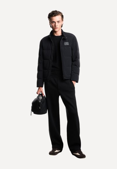 Zwarte pufferjack met een ritssluiting aan de voorkant, een gestructureerd zwart sweater en losse zwarte broek. Houdt een kleine zwarte tas vast met een gestructureerd patroon.