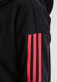 Sweat à capuche noir à texture lisse, avec trois bandes rouges verticales sur la manche gauche et une coupe décontractée.