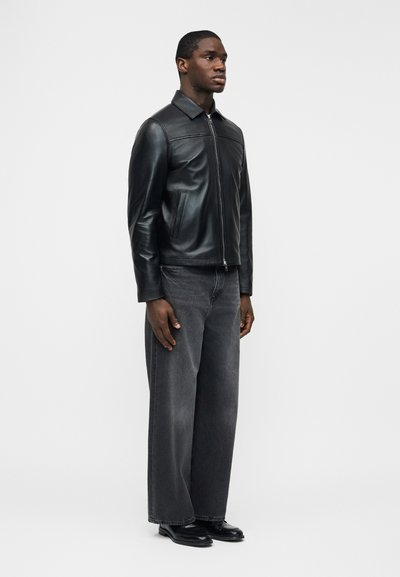 Άνδρας που στέκεται στραμμένος προς τα δεξιά, φοράει μαύρο δερμάτινο μπουφάν με φερμουάρ, μαύρο ξεθωριασμένο τζιν με φαρδιά μπότα και μαύρα παπούτσια, πάνω σε λευκό φόντο.