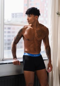 Schwarze Boxershorts mit einem blauen Bund, versehen mit dem Text "denim bros est. 89 cal", getragen von einem oberkörperfreien Model, das Tätowierungen präsentiert.