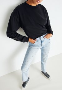 Svart långärmad sweatshirt med rund halsringning. Ljusblå jeans med dekorativa nitar längs benen. Grå sneakers fulländar outfiten.