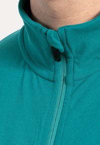 Türkise Zip-Jacke mit hohem Kragen, die eine glatte Textur aufweist und ein schwarzes Reißverschlussdetail an der Vorderseite hat. Die Nähte sind sauber verarbeitet.