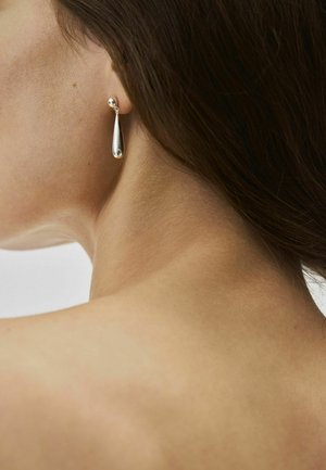 Prim-plan al gâtului și urechii unei femei purtând o cercească elegantă argintie în formă de picătură, pe un fundal simplu.