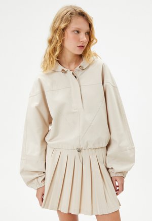 Veste beige avec capuche, fermeture à bouton, et poignets froncés, associée à une jupe plissée de la même couleur. Texture lisse dans l'ensemble.