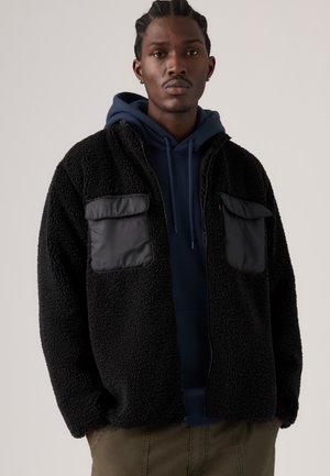 Mann mit geflochtenem Haar trägt eine schwarze Fleecejacke mit großen Taschen über einem marineblauen Hoodie und olivgrünen Hosen, steht vor einem einfarbigen Hintergrund.