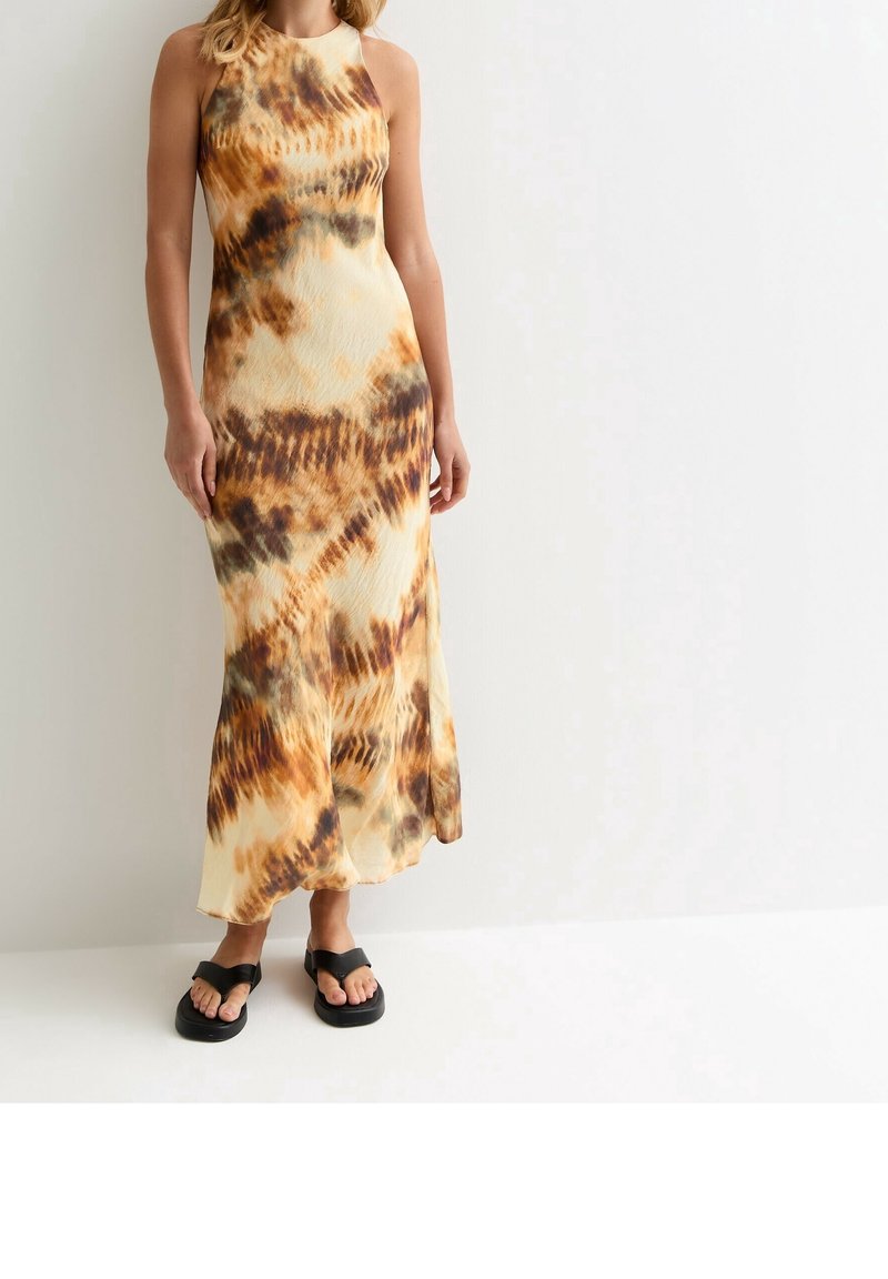 Femme portant une robe sans manches longue jusqu'aux chevilles avec un motif tie-dye marron et beige et des sandales plateformes noires, debout contre un mur blanc.