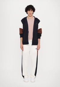 Navy and white color-block jacket, pink t-shirt, cream joggers with navy and brown accents, and white sneakers. Plain background.