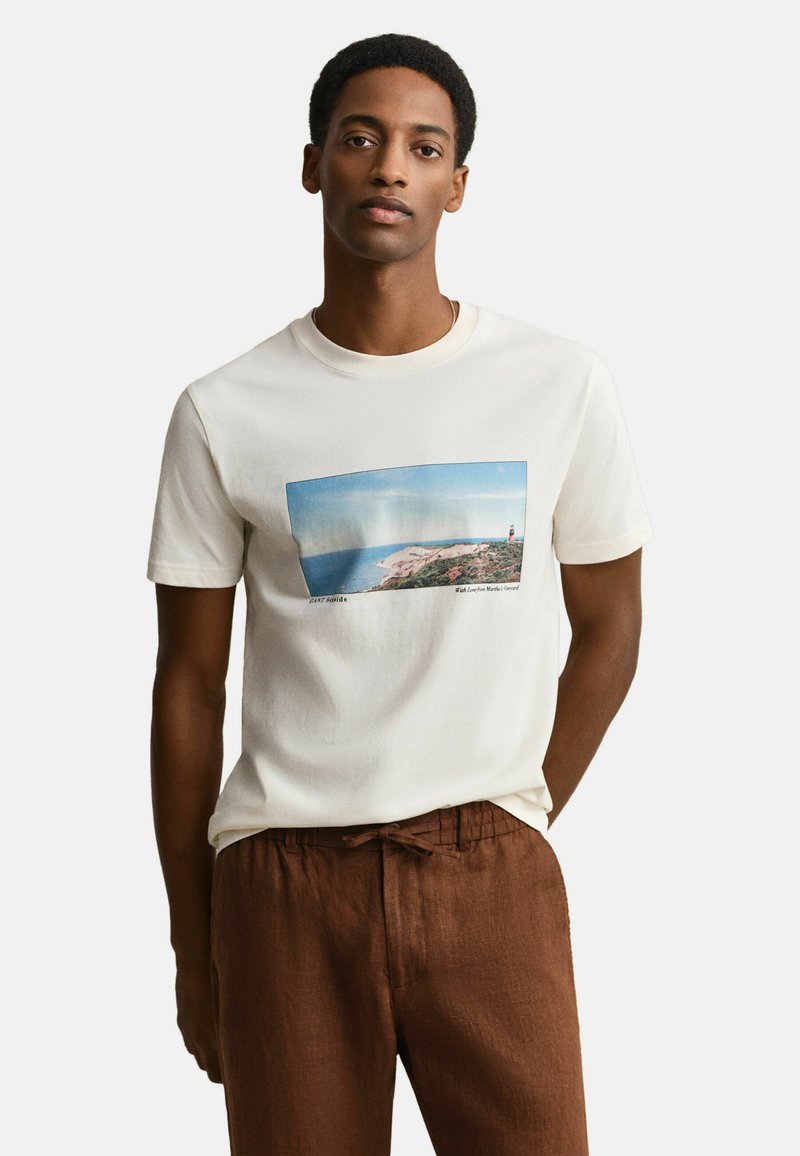 Homme portant un t-shirt blanc avec un imprimé de paysage côtier et un phare, associé à un pantalon marron à cordon, debout devant un fond uni.