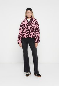Puszysty, oversize'owy sweter w ciemnobrązowym kolorze z dużymi różowymi wzorami kwiatowymi. Zestawiony z czarnymi spodniami dzwonami i czarnymi trampkami z brązowymi akcentami.