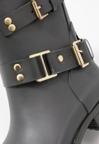 Botte en caoutchouc grise avec quincaillerie dorée, incluant des boucles et des clous, avec une texture lisse et un design légèrement courbé.