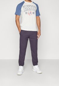 Miestenské tričko s krátkym rukávom v bielej a modrej farbe s grafikou Yankees, skombinované s tmavofialovými jogger nohavicami a bielymi športovými topánkami.