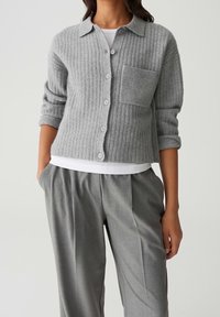 Cardigan gris côtelé avec boutons sur le devant, un col et une poche poitrine, associé à un pantalon gris ajusté et un sous-vêtement blanc.