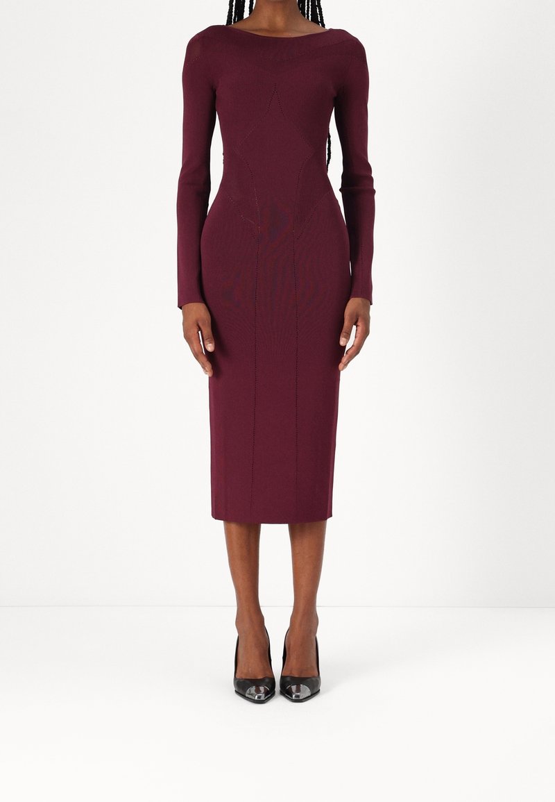 Femme portant une robe bordeaux ajustée à manches longues, longueur genou, avec des motifs texturés subtils et des talons hauts noirs pointus.