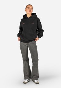Mulher vestindo um hoodie preto com texto vermelho, jeans cinzas com pés de flanela e tênis brancos com listras pretas, de pé contra um fundo branco.
