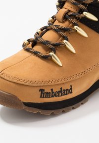 Bota de cuero marrón con acentos negros, herrajes dorados y sistema de cordones texturizados. Presenta un prominente logo de "Timberland" cosido en el lateral.