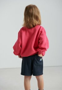 Czerwona bluza z dużymi, szerokimi rękawami, zestawiona z ciemnogranatowymi, futrzanymi szortami z kieszenią oraz zieloną, białą i niebieską metką.