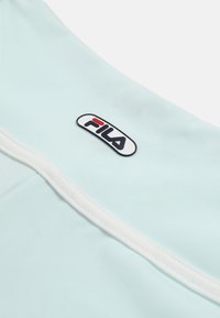 Prenda azul claro con ribete blanco que presenta un pequeño parche con el logo de FILA en negro, rojo y blanco.