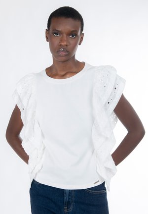 Schwarze Frau mit kurzen Haaren, die ein weißes, ärmelloses Top mit Rüschen und blaue Jeans trägt. Sie steht mit den Händen hinter dem Rücken vor einem weißen Hintergrund.