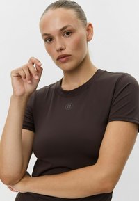 Brązowa dopasowana koszulka z krótkim rękawem o okrągłym dekolcie i subtelnym logo na środku, wykonana z gładkiego, elastycznego materiału.