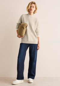 Hellgrauer Strickpullover mit Texturdetails und einem hohen Kragen, kombiniert mit dunkelblauen, weit geschnittenen Jeans und weißen Turnschuhen, während er eine tannfarbene Steppjacke hält.