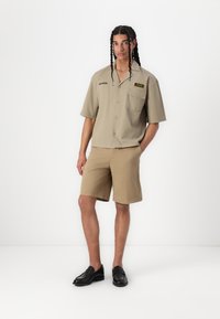 Camicia a maniche corte marrone chiaro con tasca a toppa e etichetta nera, abbinata a pantaloni corti beige e mocassini neri, indossata da un modello su uno sfondo bianco.