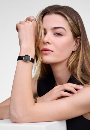 Montre à bracelet en cuir noir avec un cadran carré rehaussé d'or. Le modèle porte des bagues superposées et un bracelet, avec des cheveux châtain clair et un maquillage neutre.